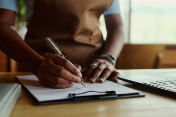 Naklejka na ściany i meble African American woman entrepreneur writing notes on clipboard near laptop representing small business management. Image suitable for accounting, retail planning and marketing