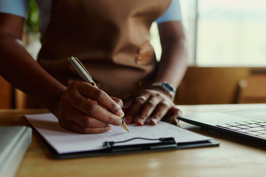 African American woman entrepreneur writing notes on clipboard near laptop representing small business management. Image suitable for accounting, retail planning and marketing