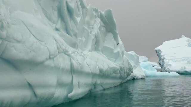 Massive Iceberg Wall Framing a Narrow Water Channel. Close cold-water view follows a tall textured blue-white iceberg edge beside calm dark water, with additional ice formations visible farther back.