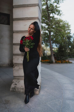 Stylish woman in black dress holding red peony bouquet beside stone pillar