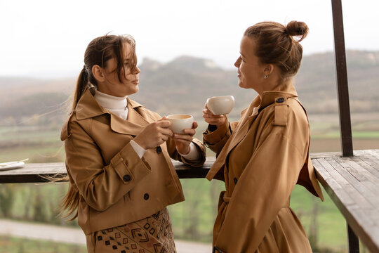 Morning coffee conversation between two women on a scenic balcony overlooking rolling green hills and landscape