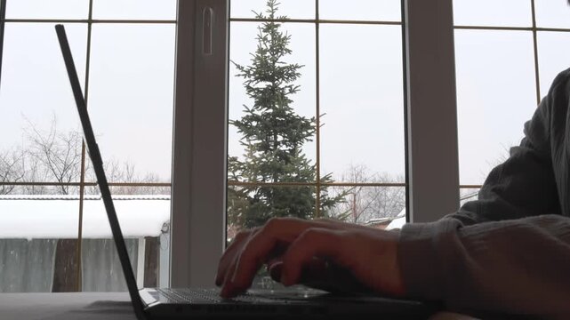 A man is working on a laptop, quickly tapping the keyboard keys while sitting at a table by the window on a cold winter day