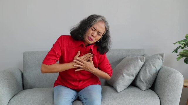 Senior Asian woman experiencing sudden chest pain while sitting on a sofa at home, representing heart disease, health problems, and medical emergency.