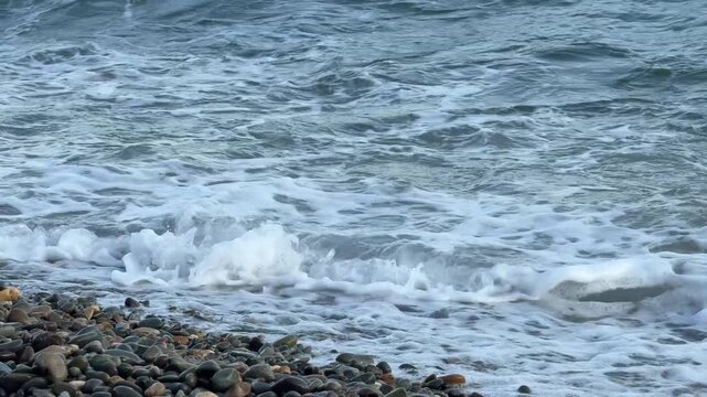 Pebble beach shoreline with frothy blue water and rolling tides crashing ashore. Static shot.