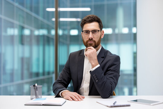 A portrait of a young businessman in a suit and glasses, sitting at an office desk, resting his hand on his beard and looking thoughtfully at the camera