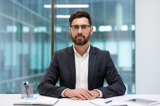 A portrait of a young businessman in a suit and glasses, sitting at an office desk with his hands clasped, looking confidently at the camera
