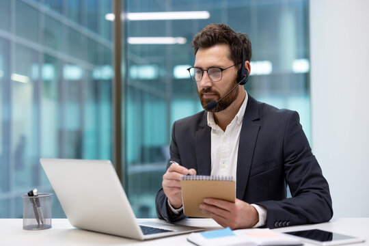A young man in a suit and headset sits at a desk in an office, looking at a laptop screen and taking notes in a notebook