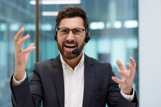 A close-up portrait of a young man in a suit and headset sitting in an office, shouting emotionally and furiously waving his hands at the camera