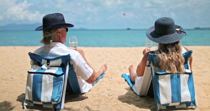 Senior couple relaxing in beach chairs on sunny Ko Samui beach, man and woman raising glasses of white wine, enjoying luxury resort vacation and celebrating retirement. Travel to Thailand. Back view
