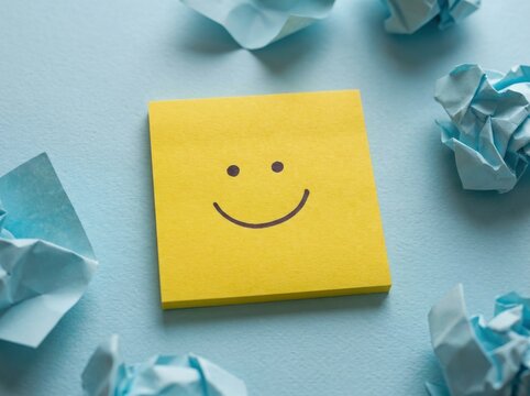 A bright yellow square sticky note featuring a simple black hand-drawn smiley face sits centered on a textured light blue background surrounded by crumpled blue paper scraps