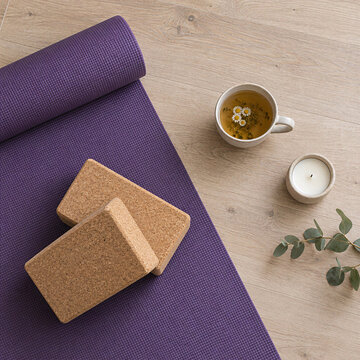 Purple yoga mat with cork blocks, cup of chamomile tea, candle, eucalyptus on wooden floor, top view