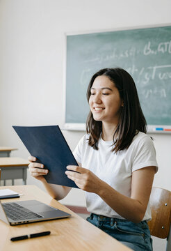 Education and academic success, happy student reading a report in a classroom with a laptop and chalkboard for learning and school development goals