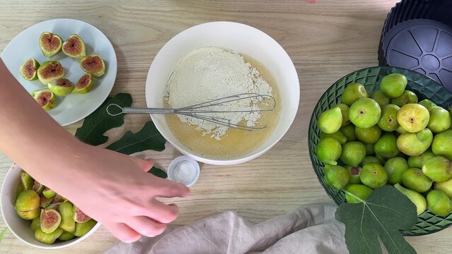 a person reaches toward a small bowl of white powder on a wooden table, surrounded by green figs in bowls and a mixing bowl with flour and a whisk