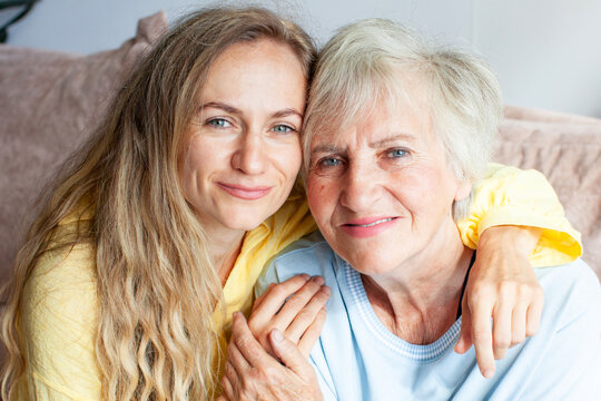 Young woman hugs elderly woman, her mom, on sofa, both smiling warmly at camera. Daughter embraces her older relative with arm around her shoulders, sharing close and affectionate family moment.