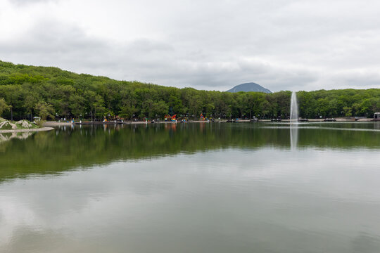 A calm park lake with a central fountain creating tall spray and clear reflections of dense green trees under an overcast sky, backed by a distant mountain and peaceful shoreline paths for walkers