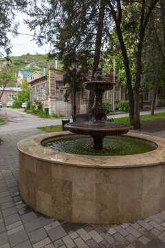 A shaded stone fountain in a quiet town park framed by tall trees, benches and historic buildings. Gentle water flow, paved walkways and leafy surroundings create a peaceful scene. Pyatigorsk, Russia