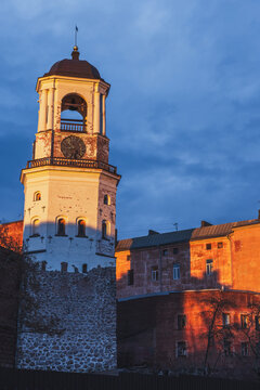 A historic clock tower of Vyborg and old town buildings glow in warm sunset light against a deep blue dusk sky, highlighting textured brickwork and an atmospheric evening mood. Vertical photo