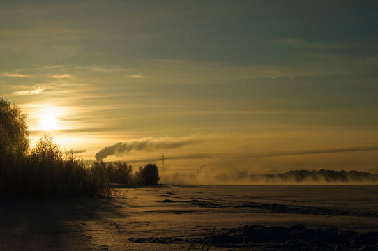 Winter landscape sunset background. A warm golden sun setting over a frozen river. Silhouetted trees and reeds on the bank with smoke rising on the horizon
