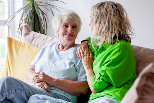 Older woman enjoys tender visit with younger woman seated close beside her. Smiling senior rests comfortably on sofa as her companion offers reassuring touch in welcoming living room.