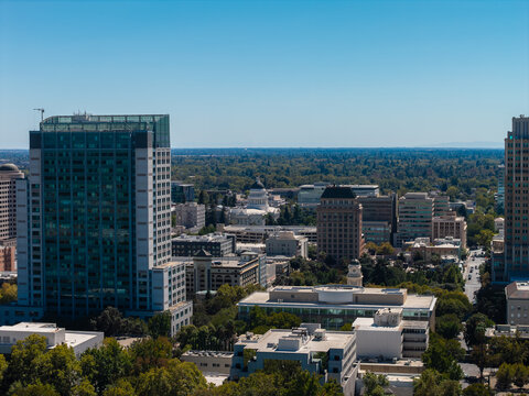 Aerial perspective of downtown Sacramento buildings and streets, showing the distant dome of the California State Capitol surrounded by dense urban tree canopy under a clear sky.