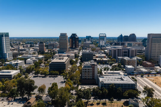 Aerial view shows the city center of Sacramento, California, displaying office towers, commercial buildings, paved streets, parking lots, and urban trees under a clear daytime sky.