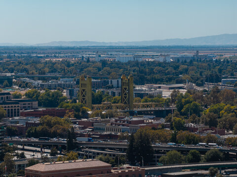 Aerial view of Tower Bridge spanning the river in Sacramento, California, showing urban architecture, highway traffic, and trees extending toward the horizon under clear daylight.