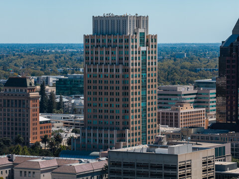 Aerial view shows prominent office skyscrapers and commercial buildings across the downtown district of Sacramento, California, set against expansive suburban tree canopy under clear daylight.