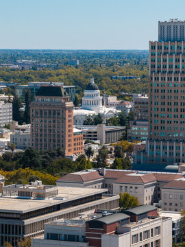 Aerial perspective reveals the dome of California State Capitol building rising amidst downtown Sacramento architecture, skyscrapers, government offices, and urban canopy on a clear sunny day.