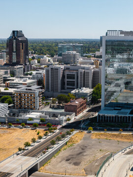Aerial view of downtown Sacramento California showing corporate skyscrapers, modern glass facades, urban roadways, vacant lots, and trees stretching towards the flat horizon on a sunny day.