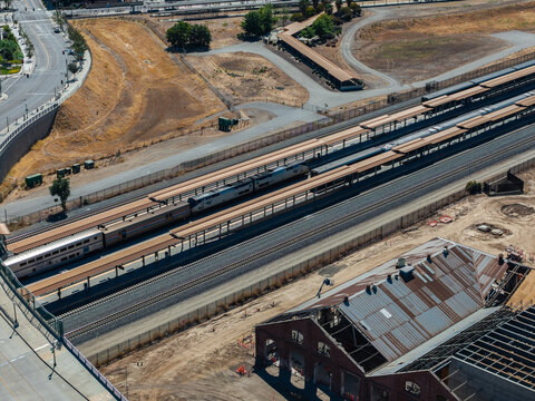 Aerial view shows passenger trains stopped alongside long boarding platforms at Sacramento Valley Station in California, with historic railroad shops undergoing renovation nearby.