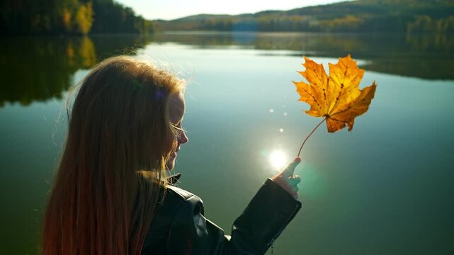 A little girl is sailing on a boat on a lake and holding an autumn leaf in her hand, swaying in the wind. On the shores of the lake there is an autumn forest in bright autumn colors.