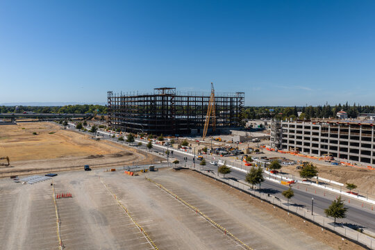Aerial view of a major construction site showing steel frame building structures, cranes, parking garage development, empty lots, and street infrastructure in Sacramento, California.
