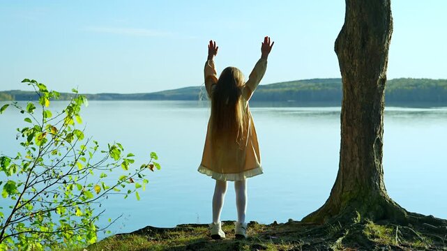 A little girl is standing on the shore of the lake and joyfully raises her arms up, admiring the landscape. A moment of childlike delight and inspiration. The harmony of nature and humanity.