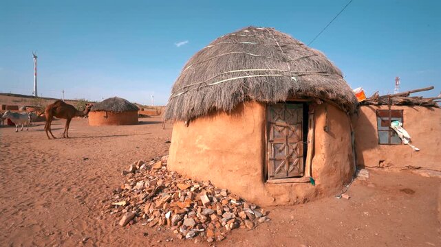 Cinematic static shot of a traditional mud hut with a thatched roof in a rural desert village with a camel walking in the background near Jaisalmer, Rajasthan, India.