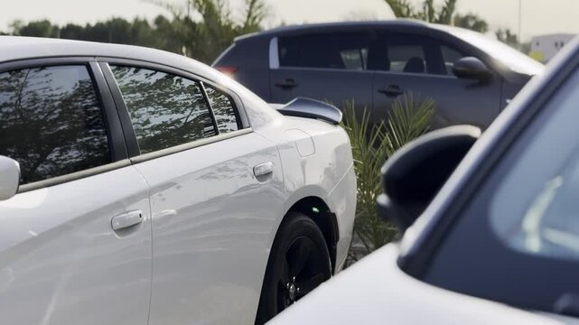 A close-up view of a modern white sedan parked in an outdoor lot during daylight. The vehicle features a rear spoiler and black wheels, sitting alongside other parked cars and green plants.