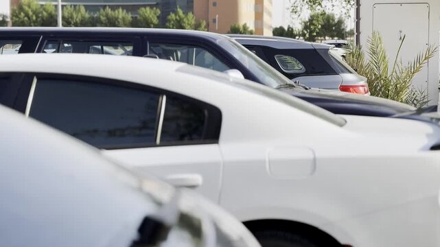 A white sports sedan sits parked in an outdoor urban lot during bright daylight. A black rear spoiler and red taillight are visible among a row of other modern vehicles and city buildings.