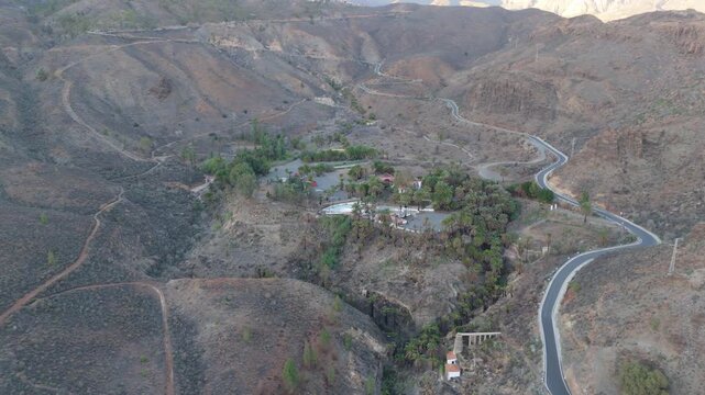 Sweeping aerial drone shot panning left over a dramatic, rugged valley on Gran Canaria. Features the hamlet of La Solana with a winding road, lush palm trees nestled among rocky hills.
