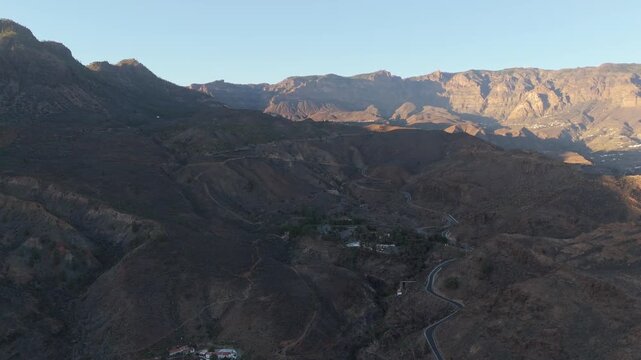 Aerial drone shot panning left over the dramatic, rugged mountain landscape of Gran Canaria. Golden hour sunlight illuminates the peaks, revealing terraced slopes and the hamlet of La Solana.
