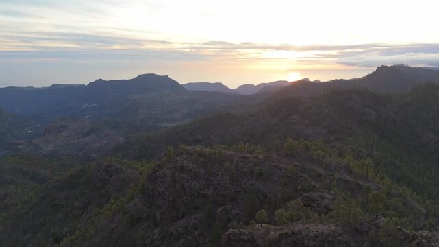 An aerial drone shot pulls back over the dramatic mountain range of Gran Canaria at sunset. The rugged pine tree landscape, reveals a vast valley and distant ocean as the sun dips below the horizon.