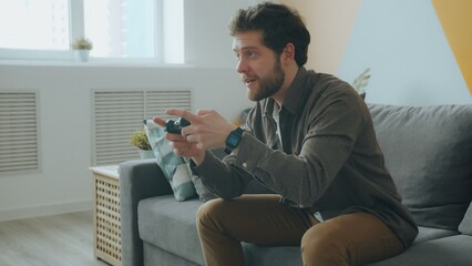 Carefree guy is playing videogame pressing joystick buttons having fun in apartment alone. Modern technology, youth and entertainment concept. © silverkblack