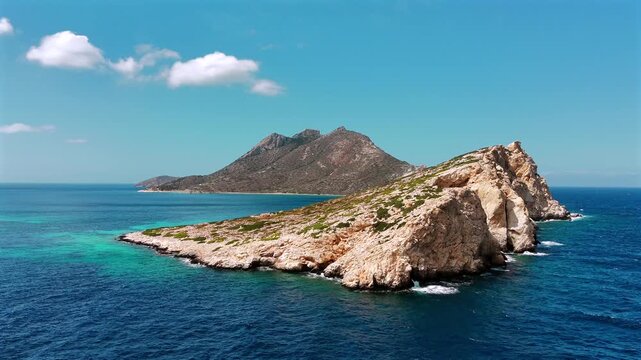 Aerial drone shot of the uninhabited rocky islet of Nikouria with its arid peak and turquoise shallows off the north coast of Amorgos, Cyclades, Greece, on a sunny day