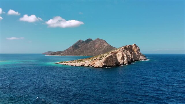 Aerial drone shot flying over the rocky promontory of Nikouria islet toward its arid summit, with clear turquoise water off the coast of Amorgos, Cyclades, Greece