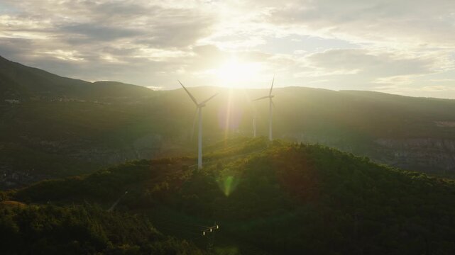 Three wind turbines stand silhouetted atop green hillside crest in Italy, blazing golden sunrise glows behind layered misty mountains above small distant lakeside village. Aerial drone view.
