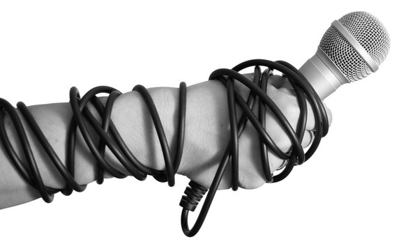 A person holds a microphone tightly wrapped in black cables on a plain transparent background. The hand grips the microphone firmly, showing a sense of control.
