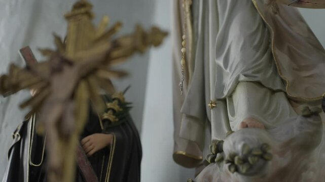 A gilded crucifix out of focus in front of a Virgin Mary figure holding a rosary, with a nun statue beside them on a chapel altar.