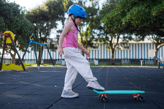 Little Girl in Blue Helmet Learning to Step on Penny Board at Outdoor Playground