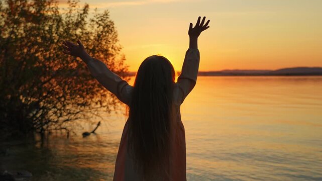 A little girl is standing on the shore of a lake at sunset and joyfully waving her arms at the golden sun. A moment of childlike delight and inspiration.