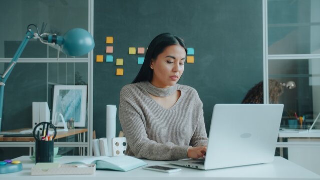 Stressed Asian woman is typing with laptop in office suffering from migraine touching head feeling headache. Health, technology and work concept.