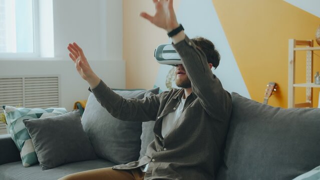 Male student in casual clothing is enjoying augmented reality glasses gesturing wearing headset at home sitting on couch alone. People and fun concept.