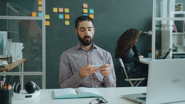 Happy Arab guy is having fun in modern office playing mobile video game on smartphone touching screen enjoying victory while coworker is working in background.